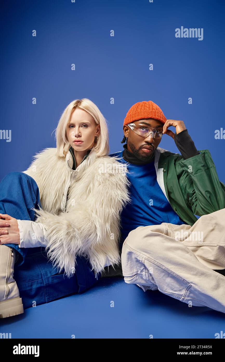 young african american man and blonde woman in winter attire sitting together on blue backdrop Stock Photo