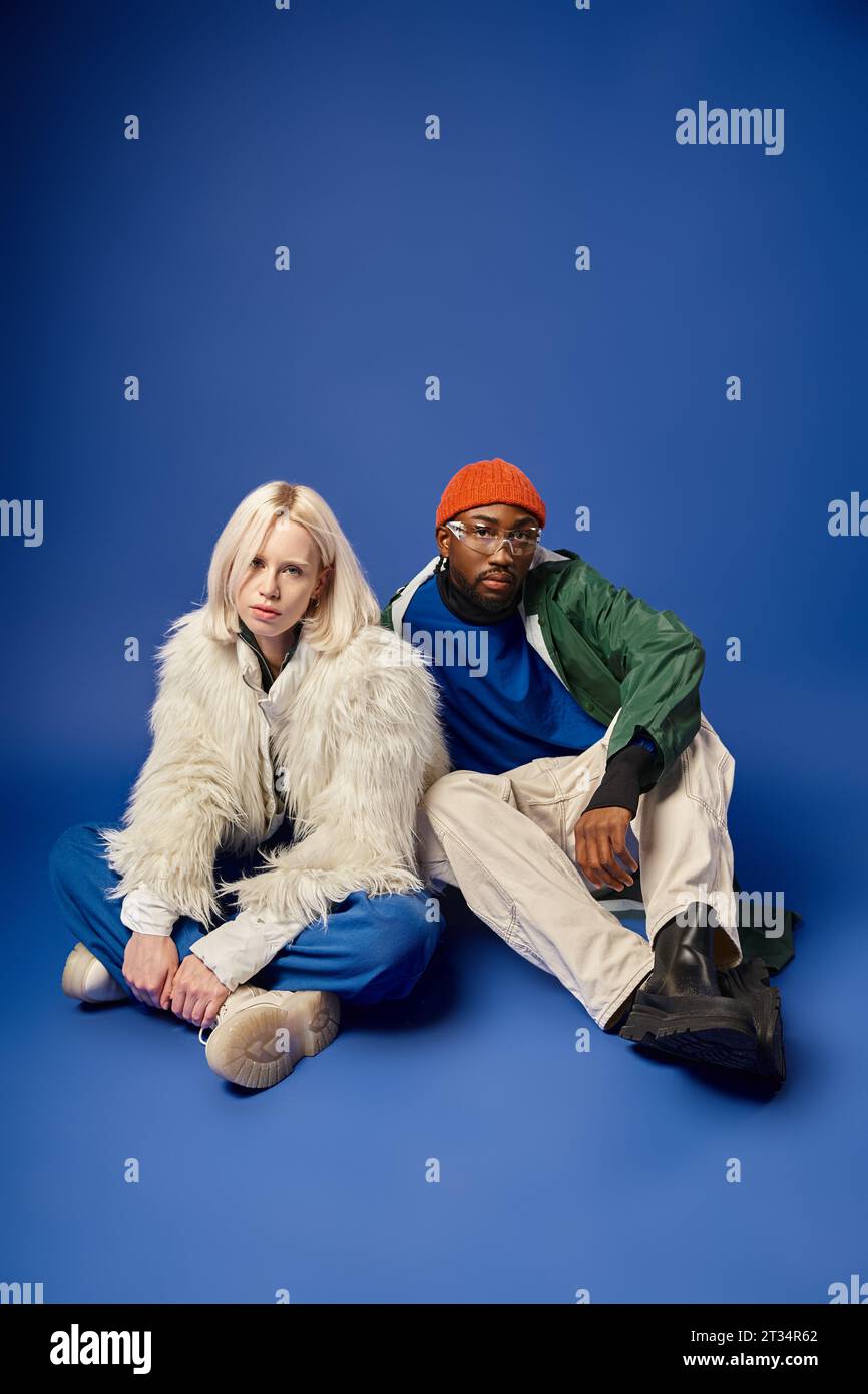 stylish african american man and blonde woman in winter attire sitting together on blue backdrop Stock Photo