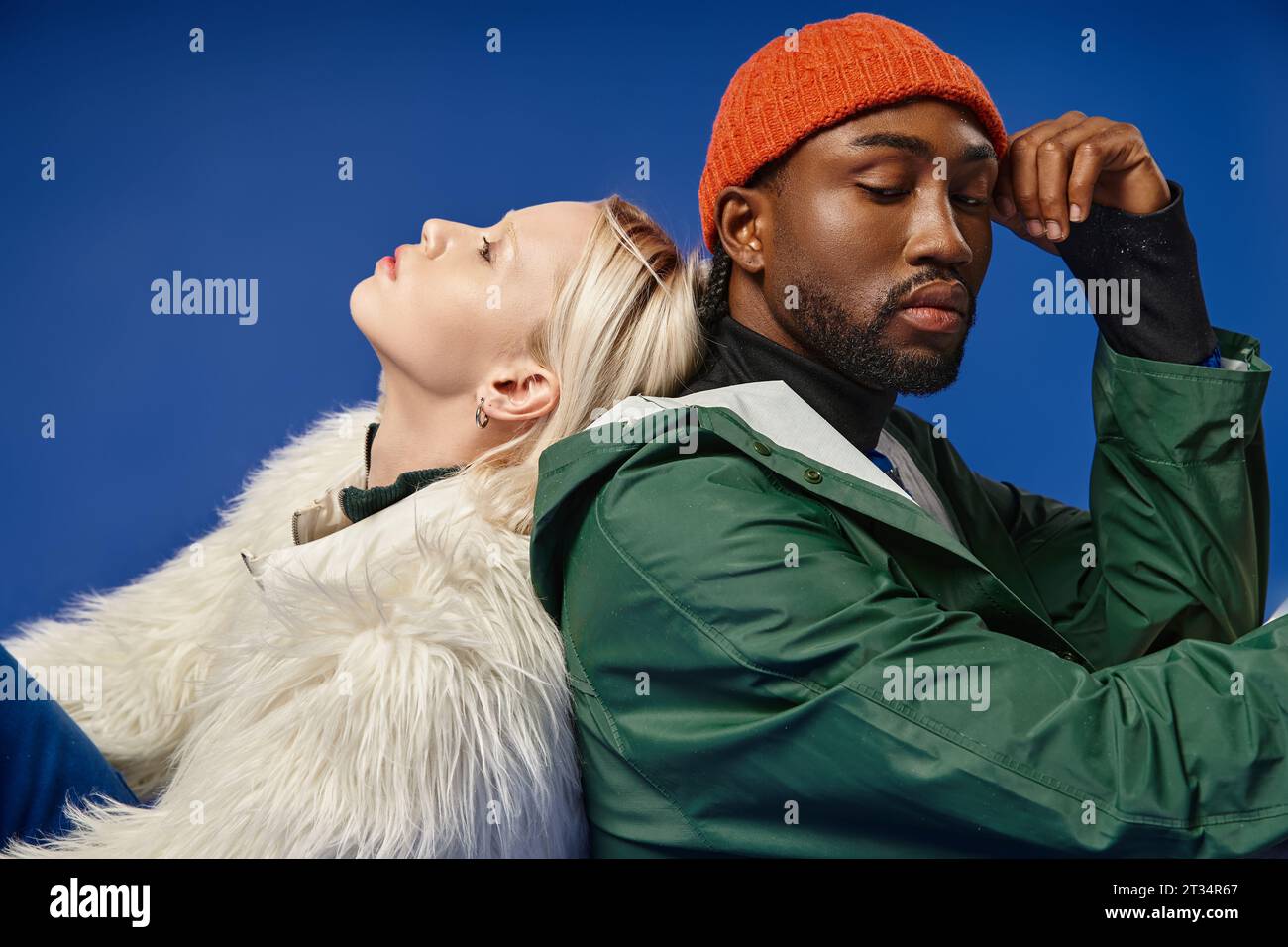 blonde woman leaning on back of african american man in hat and winter attire on blue backdrop Stock Photo