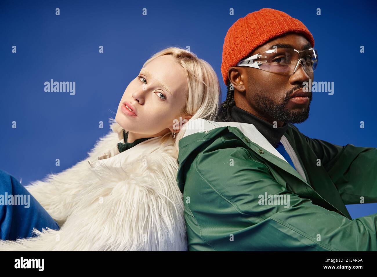 young multicultural models in winter attire sitting together on blue backdrop, diversity and fashion Stock Photo