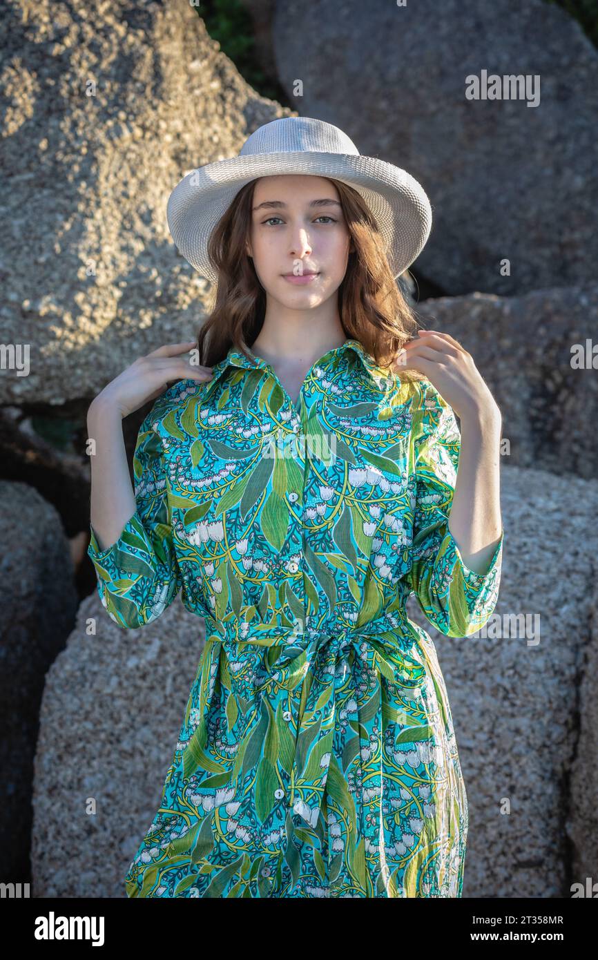 A 20-year-old Caucasian woman radiates natural beauty on the beach, adorned in a green patterned shirt dress and a hat against a backdrop of boulders Stock Photo