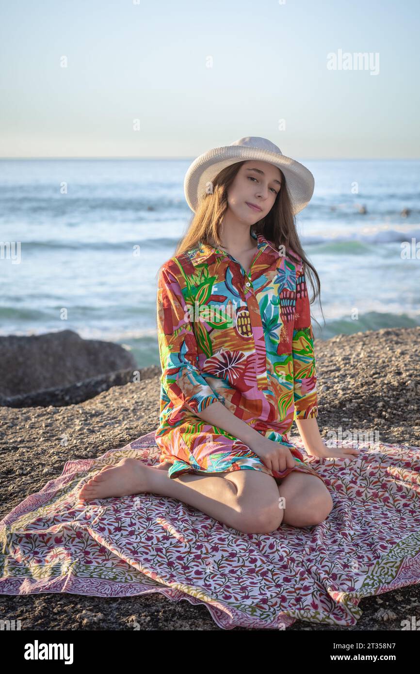 In the late afternoon glow, a 20-year-old Caucasian woman sits on a beach boulder, adorned in a vibrant, patterned shirt dress and a chic hat Stock Photo