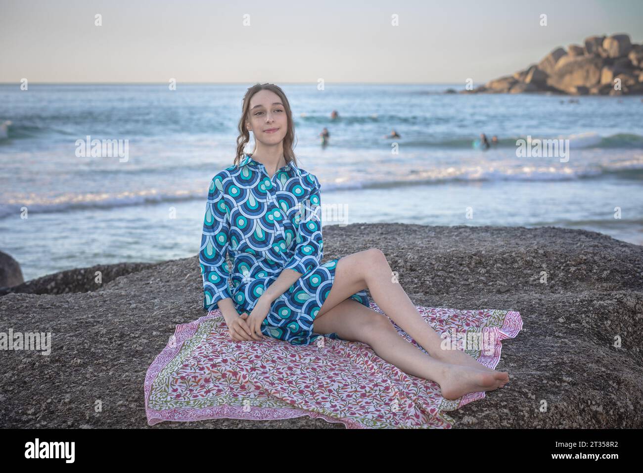 In the late afternoon glow, a 20-year-old Caucasian woman sits on a beach boulder, adorned in a vibrant, patterned shirt dress Stock Photo