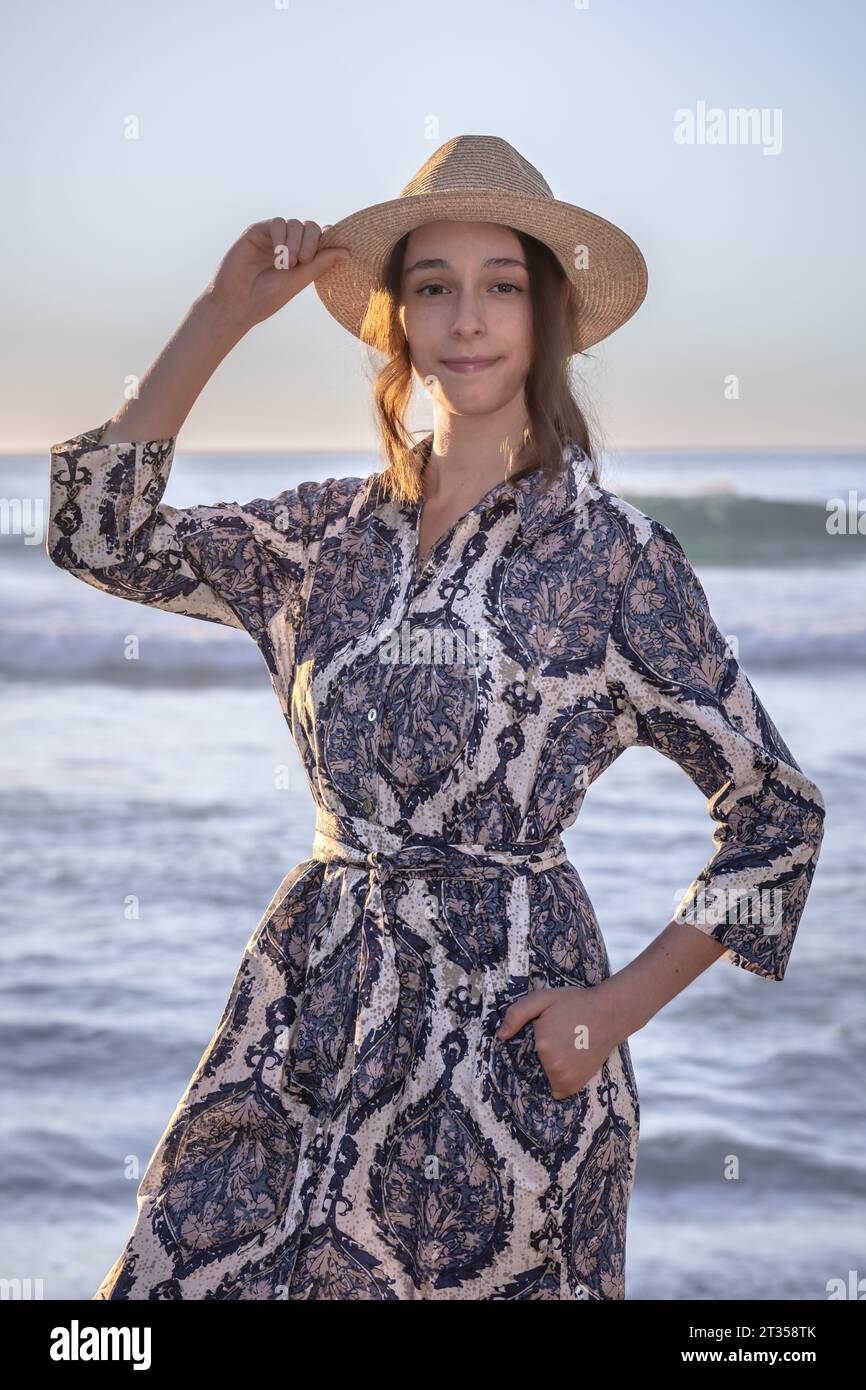 A 20-year-old Caucasian woman stands on the beach at sunset, wearing a vibrant and colorful patterned shirt dress as the sun casts a warm glow over th Stock Photo