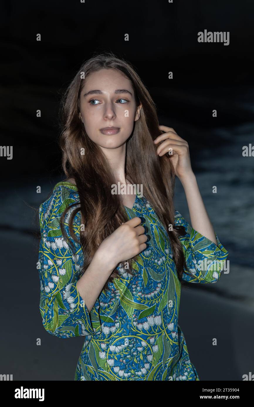 An elegant 20-year-old Caucasian woman with long flowing hair exudes grace in a close-up shot, donned in a green patterned shirt dress on the beach Stock Photo