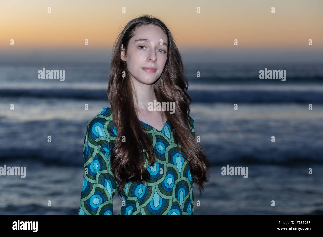 A 20-year-old Caucasian woman stands on the beach at sunset, wearing a vibrant and colorful patterned shirt dress as the sun casts a warm glow over th Stock Photo