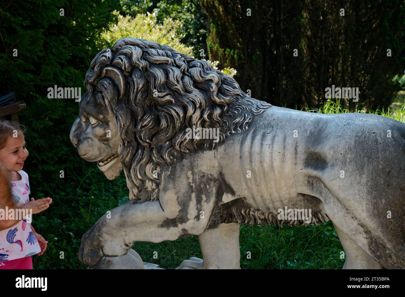 Young girl timidly interacting with the weathered mouth of a lion stone statue, set against the backdrop of the gardens of the historic Peles Castle Stock Photo