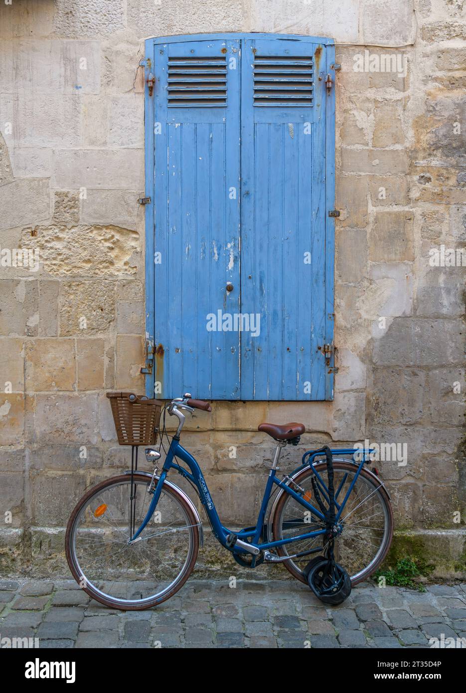 Blue bicycle with basket and crash helmet chained up, lent against stone wall with closed, blue shutters in the seaside town of La Rochelle, France. Stock Photo