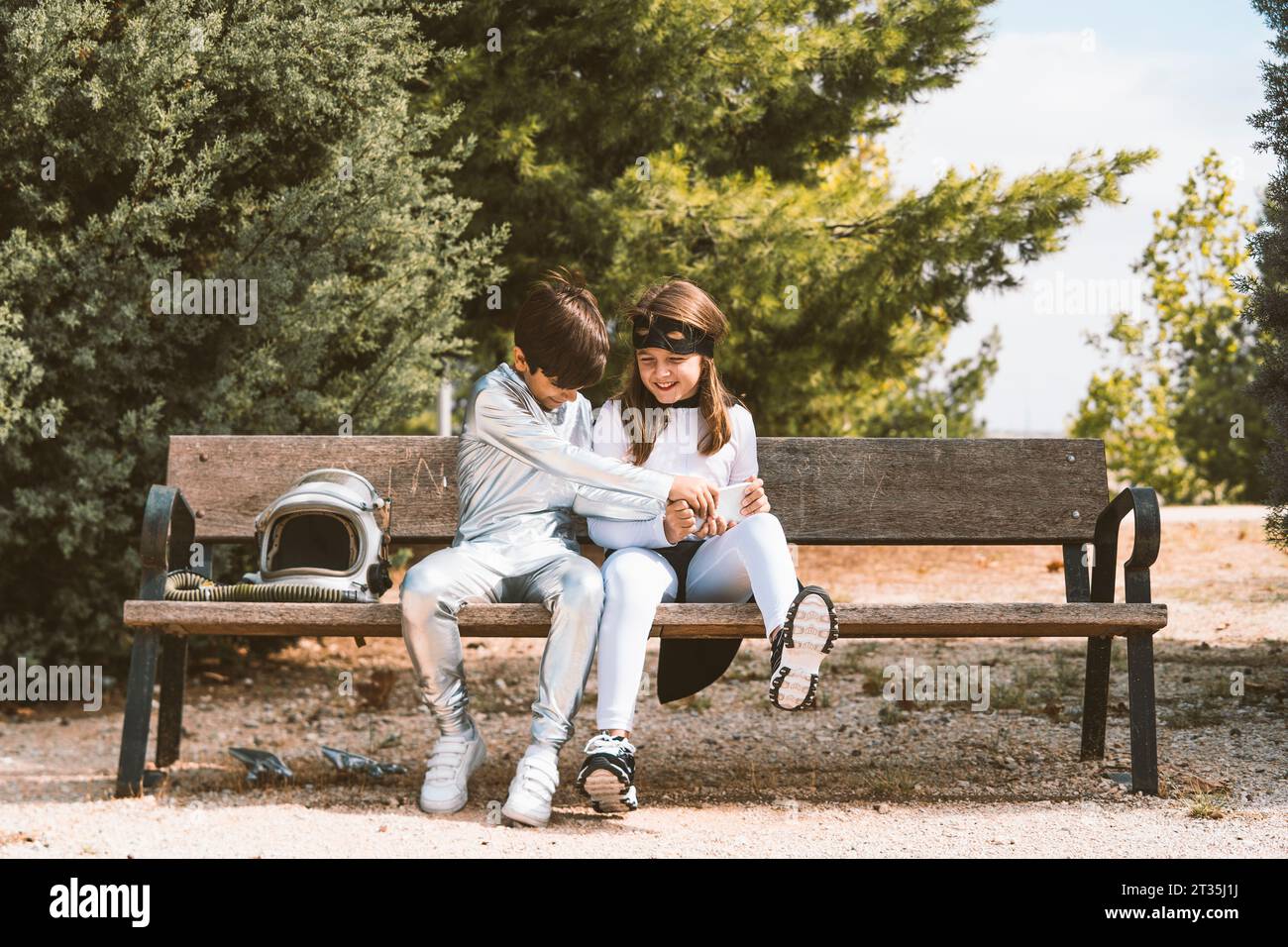 Two kids in astronaut and superhero costumes with mobile phone on park bench Stock Photo