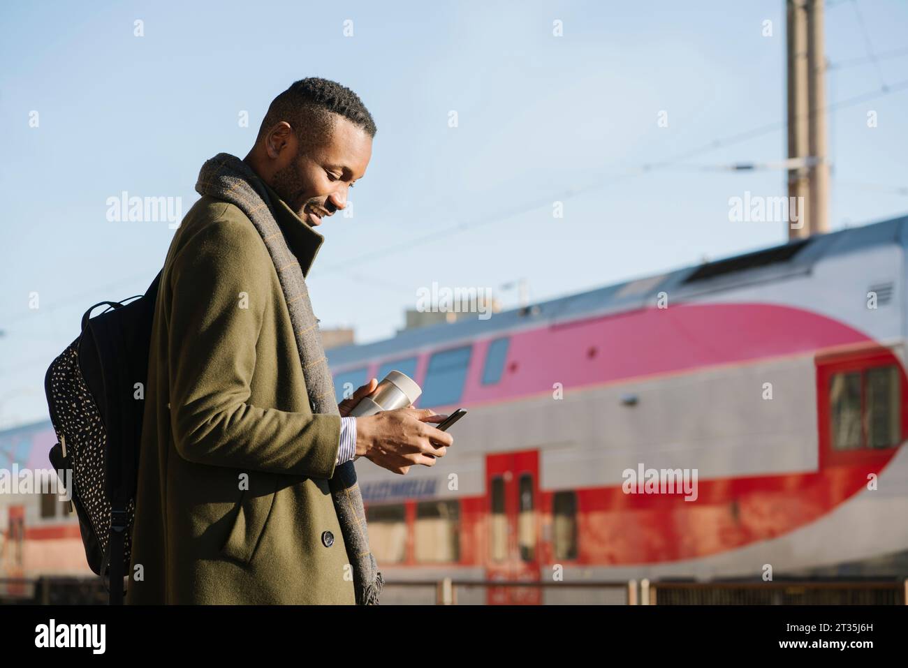 Stylish man with reusable cup using smartphone while waiting for the train Stock Photo