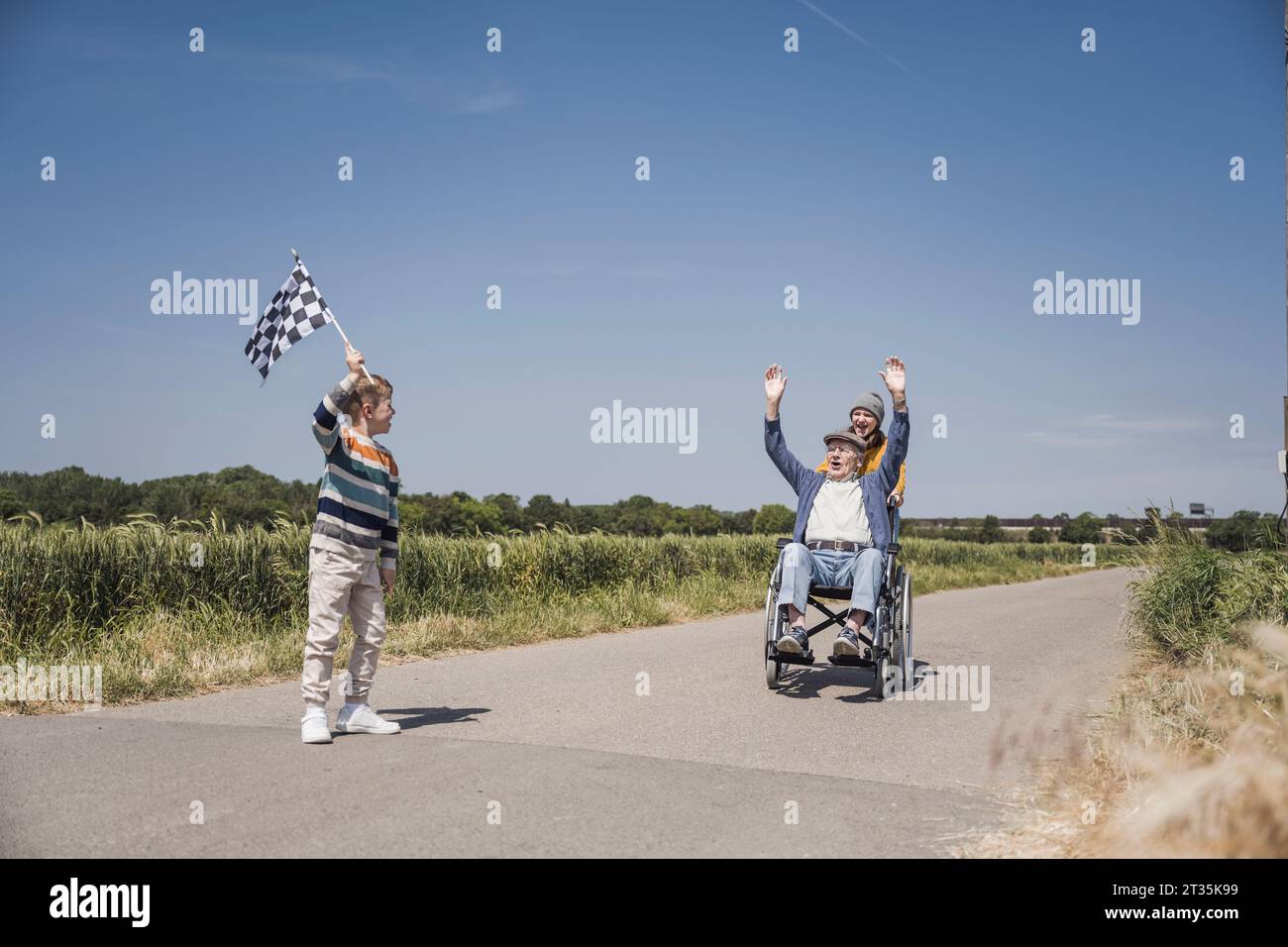 Boy holding checkered flag with senior man sitting in wheelchair on road Stock Photo
