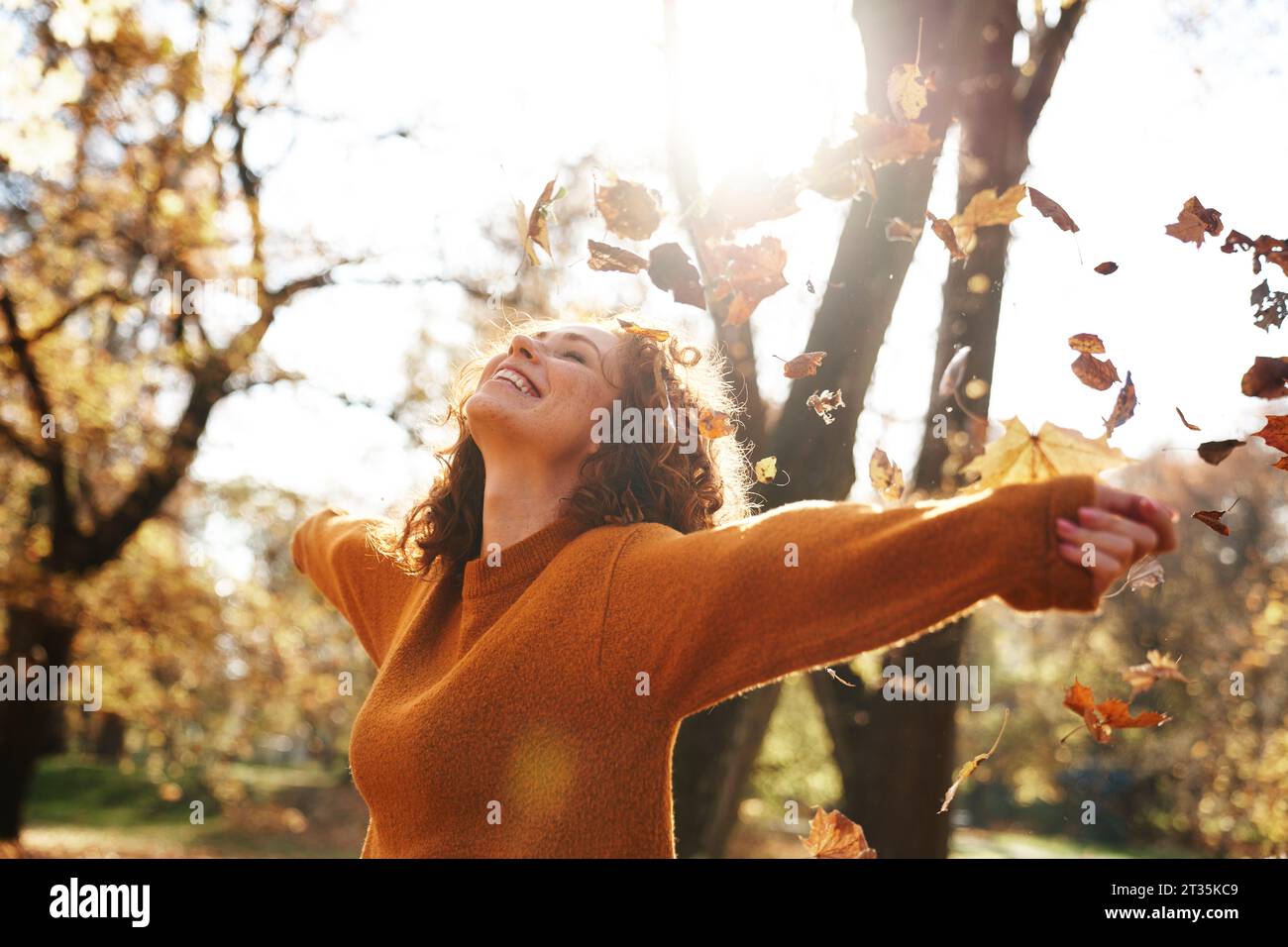 Redhead woman with arms outstretched enjoying autumn leaves falling at park Stock Photo