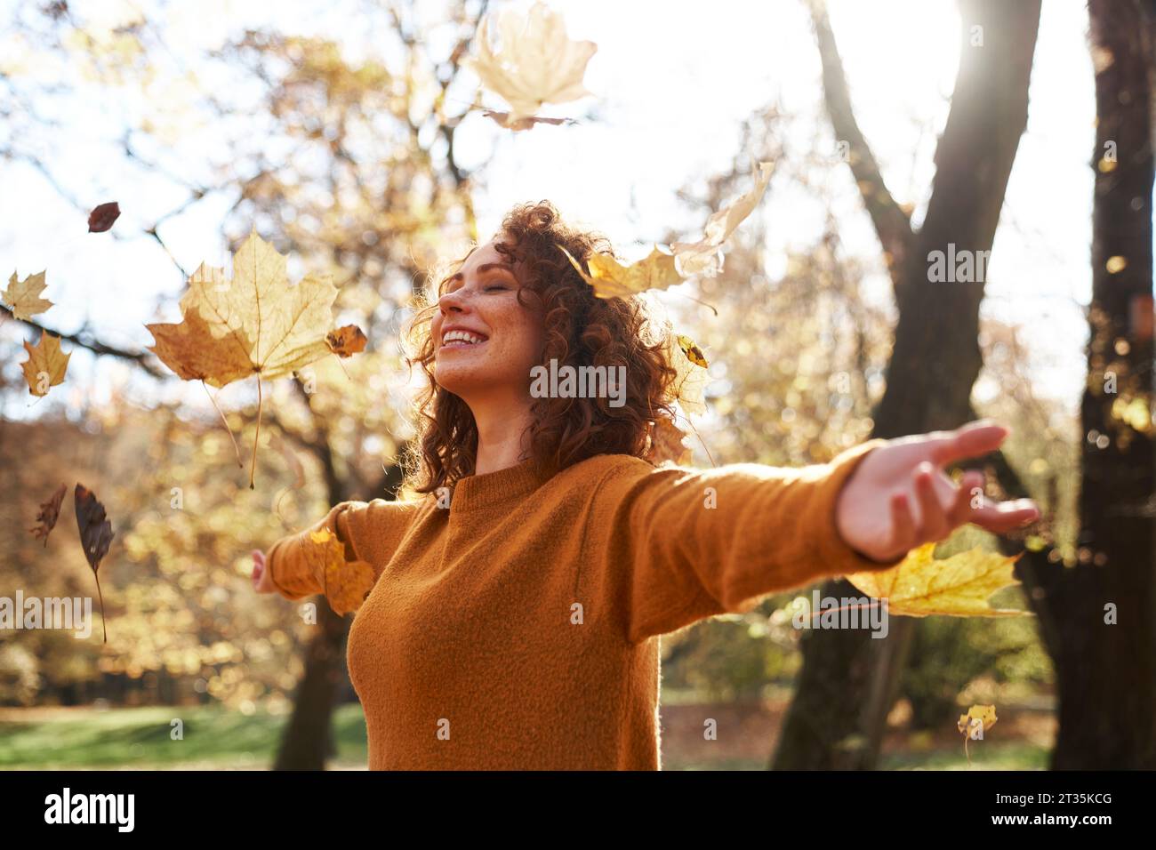 Autumn leaves falling on redhead woman with arms outstretched at park Stock Photo