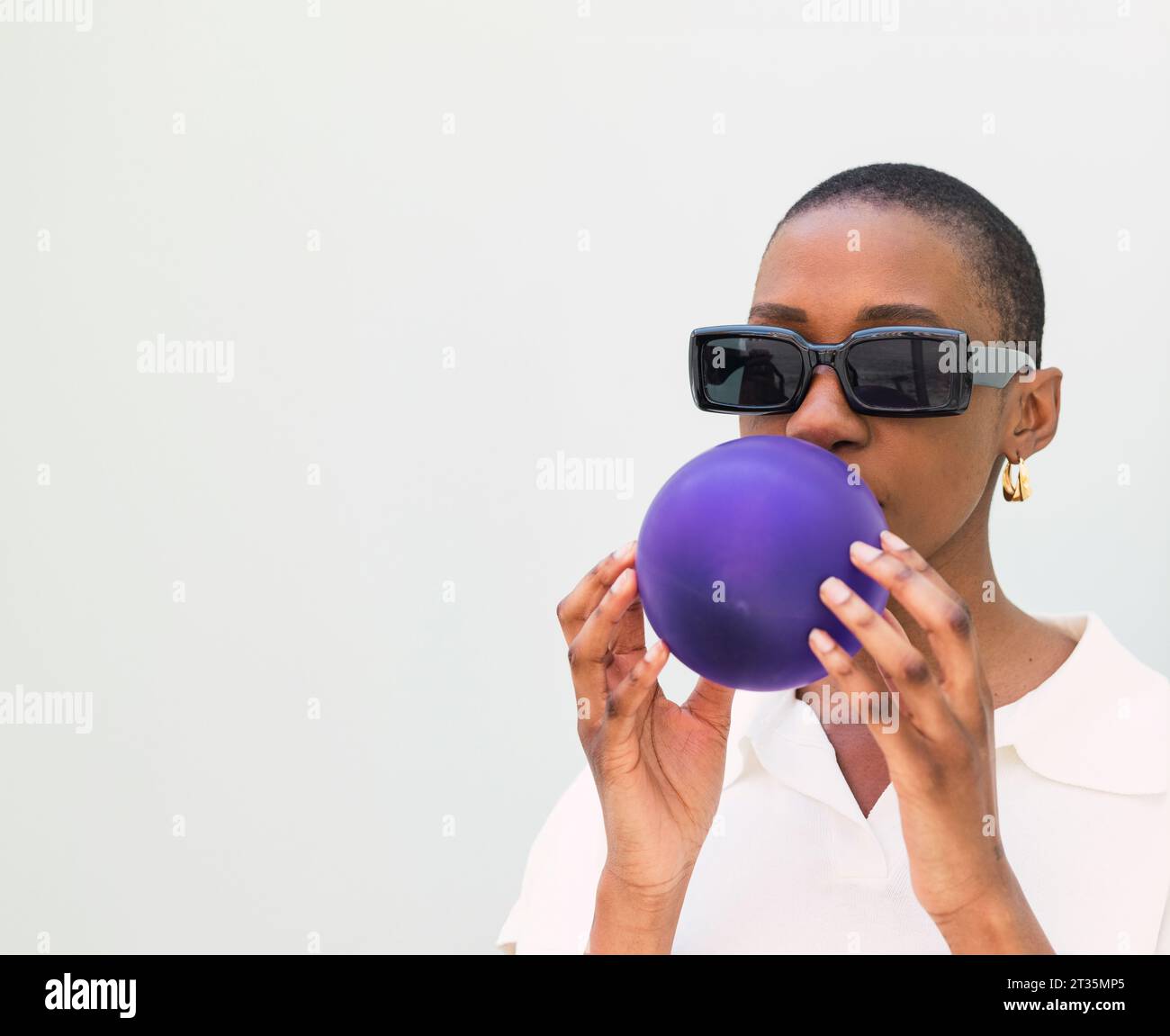 Young woman wearing sunglasses blowing balloon against white background Stock Photo