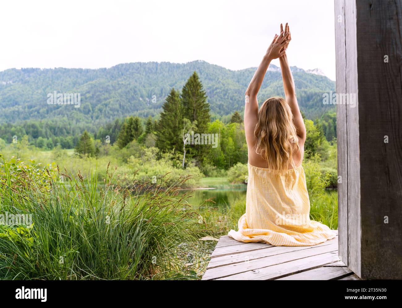 Woman with arms raised sitting on wooden boardwalk at Zelenci Nature Reserve Stock Photo