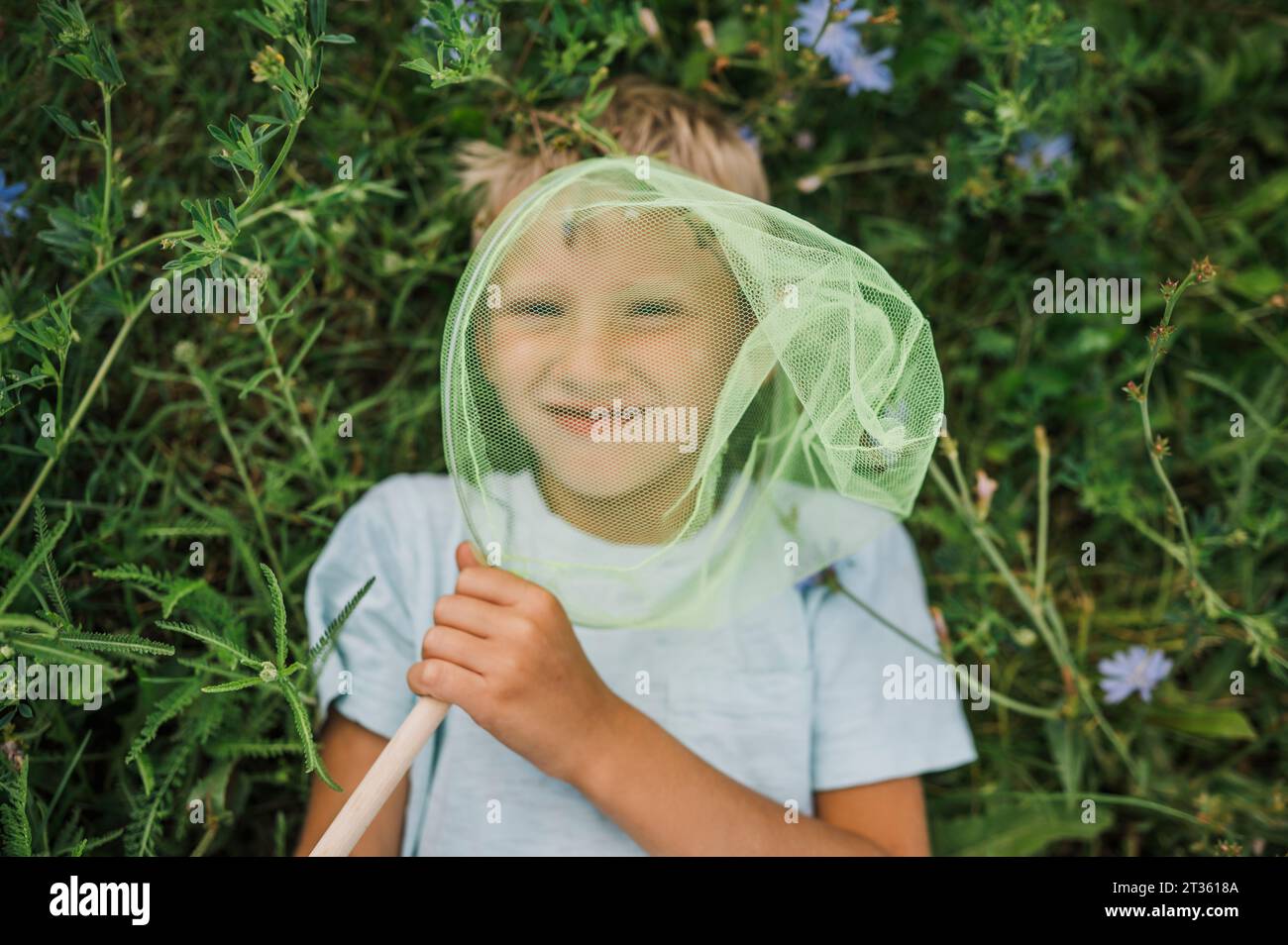 Smiling boy holding butterfly net lying down on plants Stock Photo