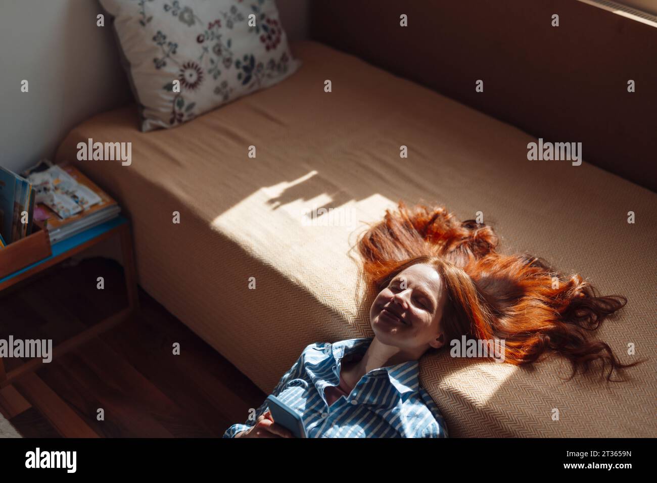 Smiling redhead woman leaning on bed with sunlight over face Stock