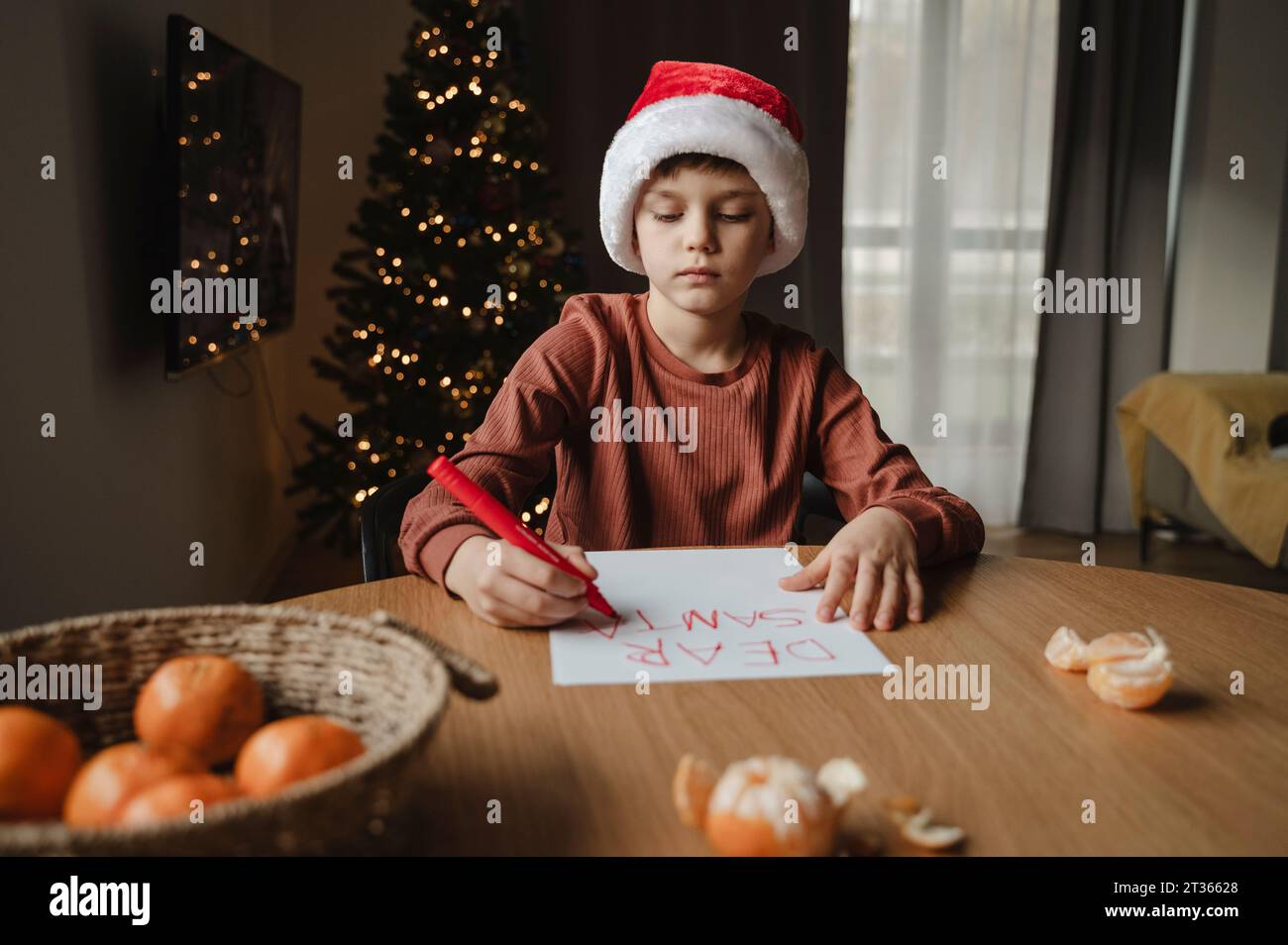 Boy wearing santa hat writing on paper at table Stock Photo