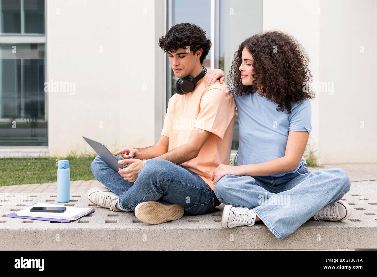 Friend Explaining Student using laptop and explaining friend on concrete bench in campus ...