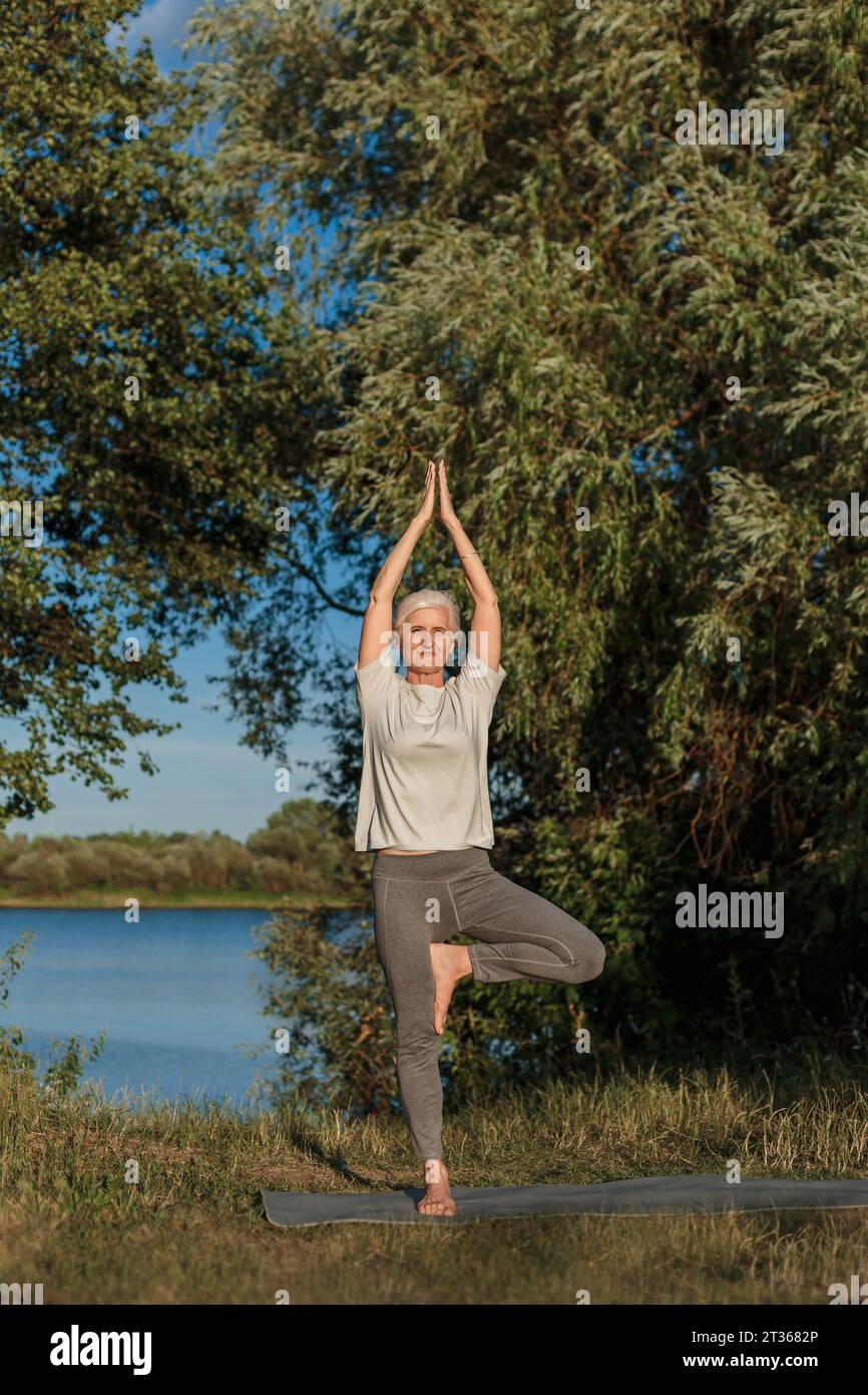 Mature woman practicing tree pose yoga on mat at lakeshore Stock Photo ...
