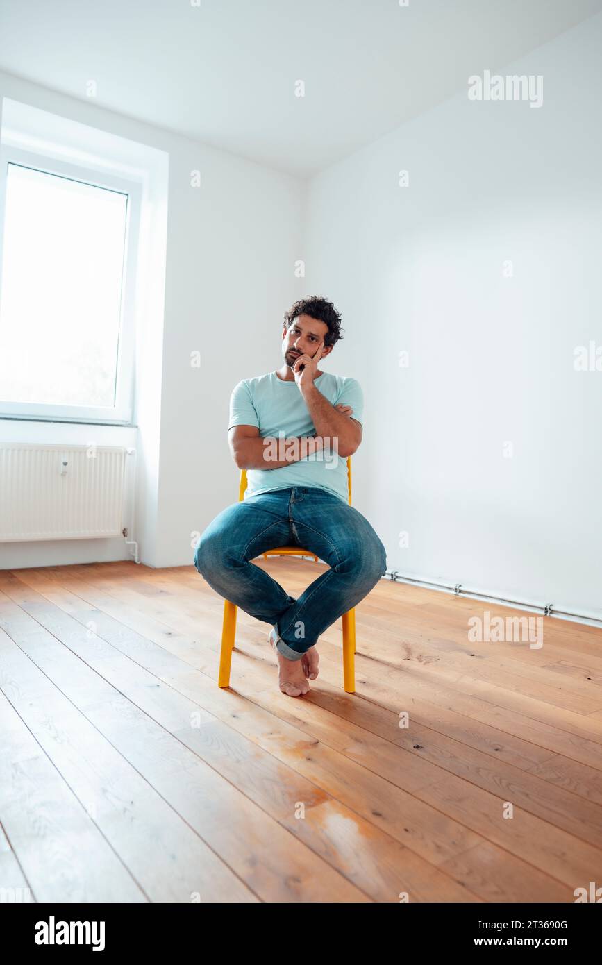 Thoughtful man sitting on chair in front of wall at home Stock Photo
