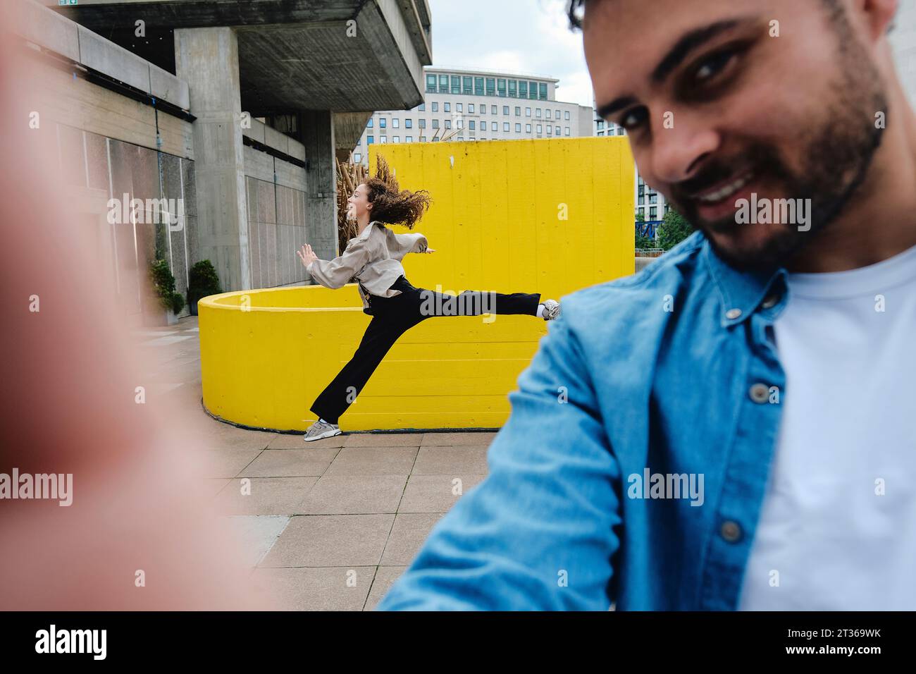 Smiling man taking selfie with woman jumping in background Stock Photo