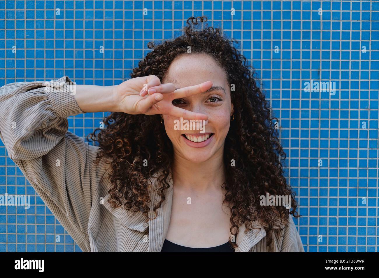 Happy woman showing peace sign in front of blue wall Stock Photo