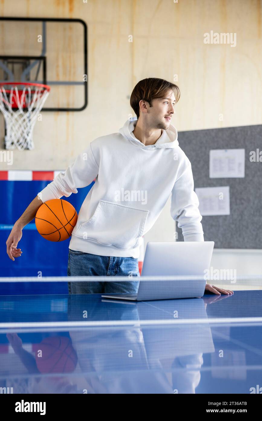 Young trainee standing with basketball and laptop in office Stock Photo