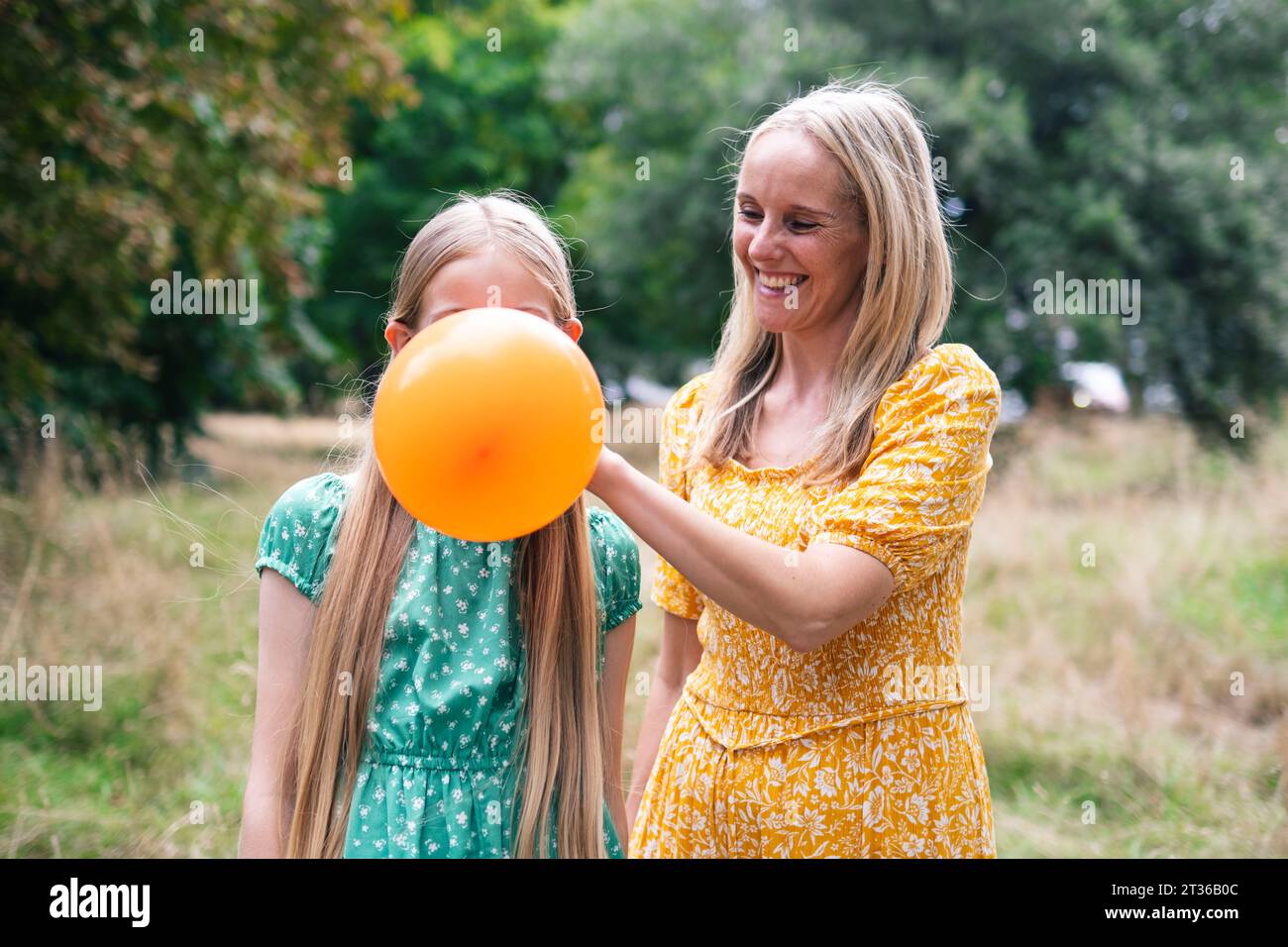 Happy woman helping daughter blowing balloon in park Stock Photo