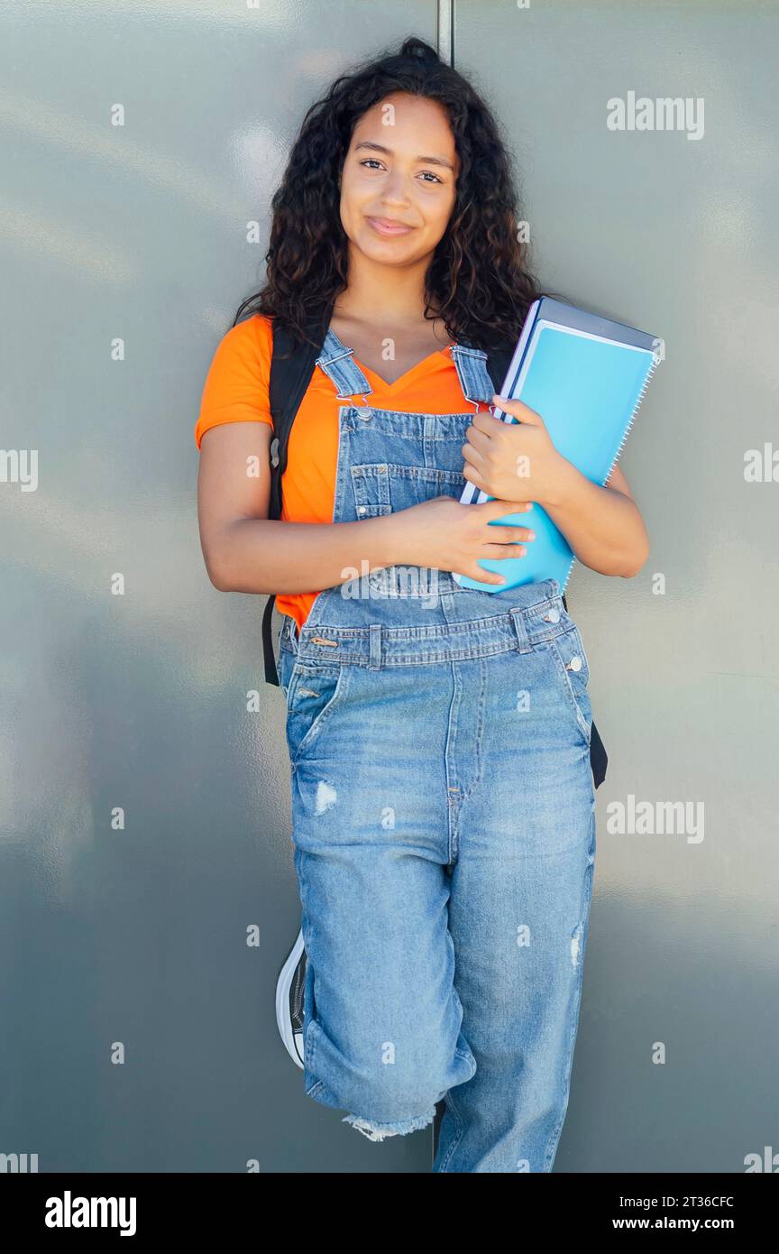 Smiling student holding spiral notebooks and standing in front of wall Stock Photo