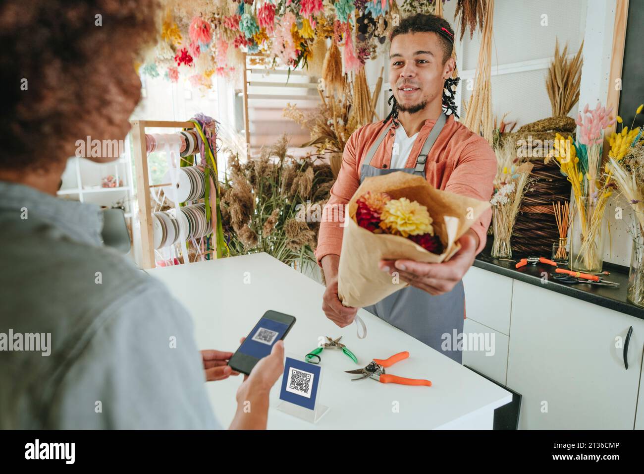 Smiling young florist giving flower bouquet to customer paying through contactless method at store Stock Photo