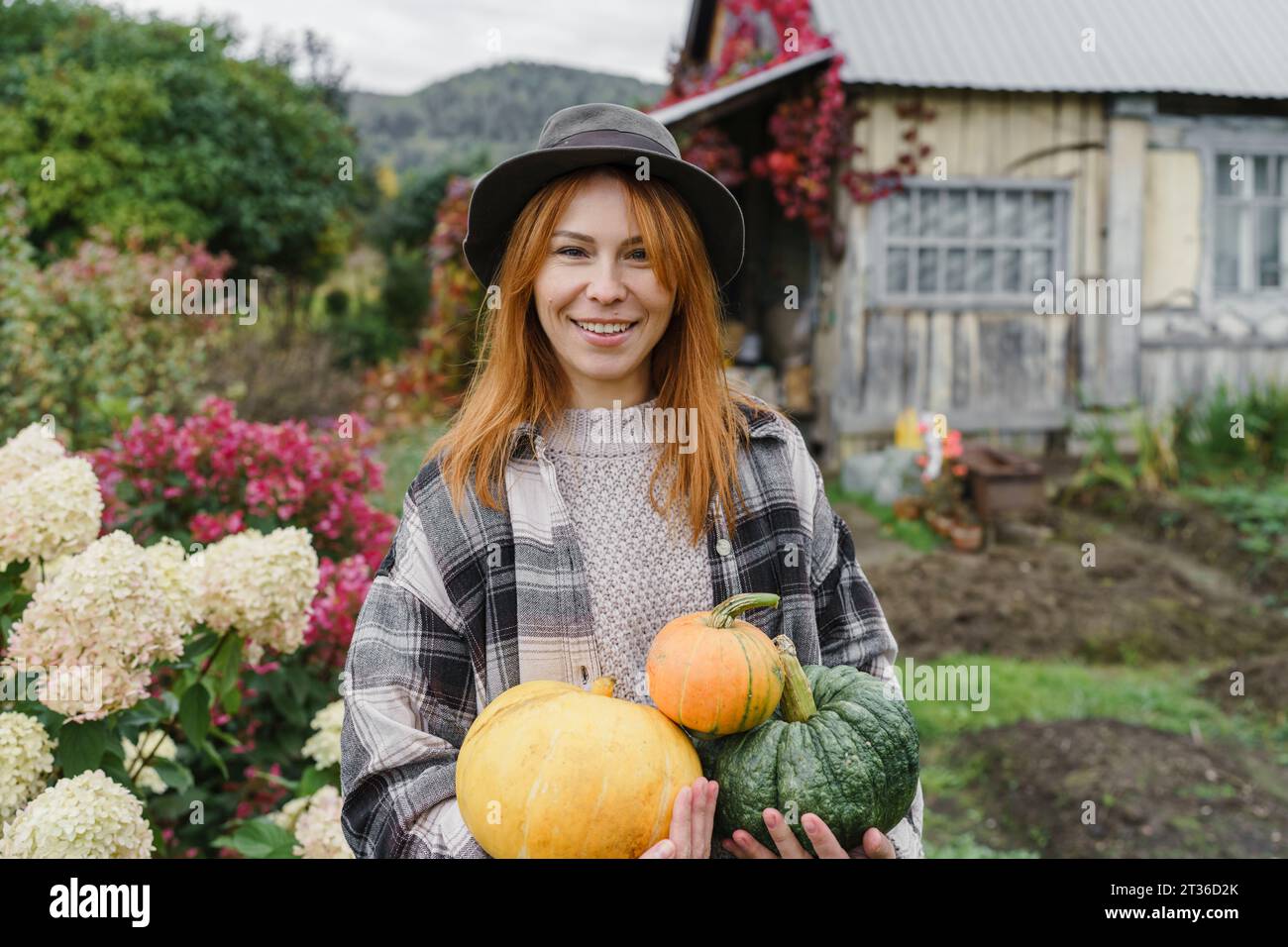 Happy redhead farmer wearing hat holding pumpkins at farm Stock Photo