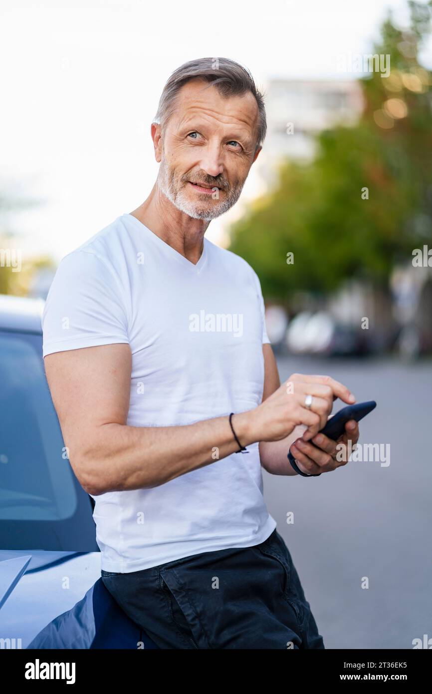 Thoughtful man with smart phone leaning on car Stock Photo