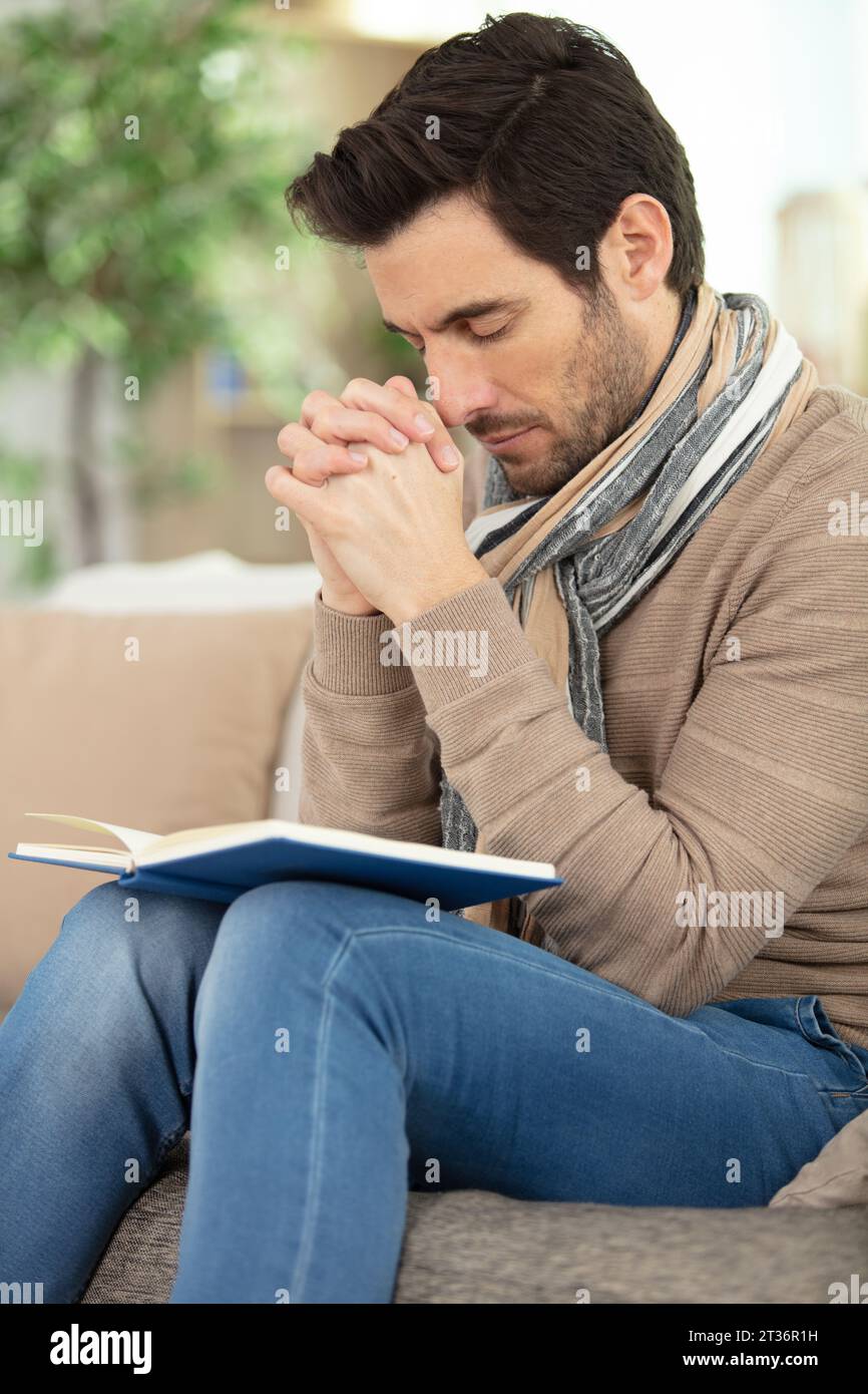 man on sofa holding book his hands in prayer position Stock Photo