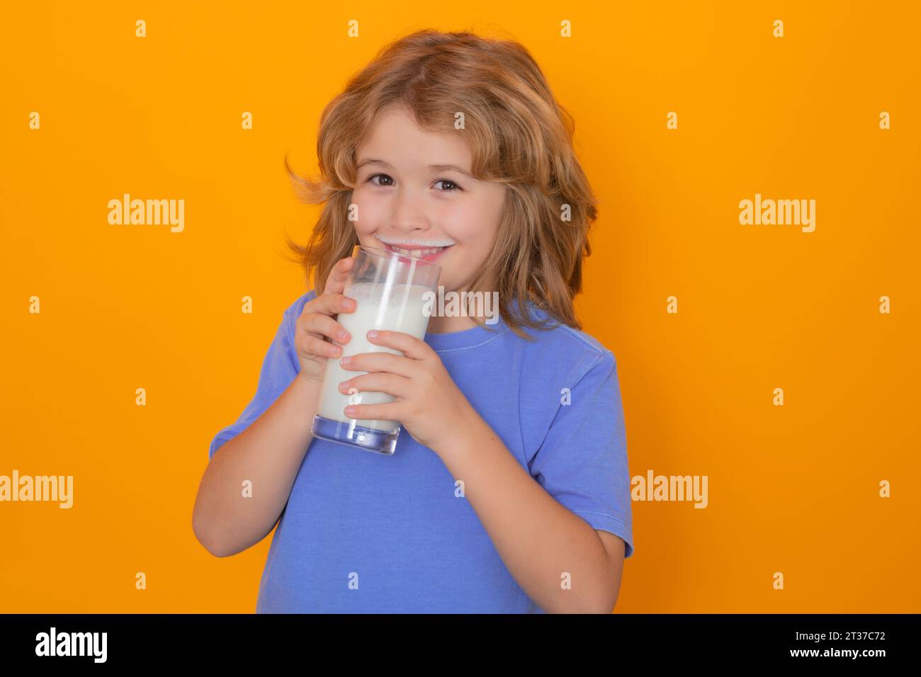 Child with a glass of milk. Cute boy in blue shirt holding glass of milk on yellow isolated studio background. Portrait of funny kid with milk mustach Stock Photo