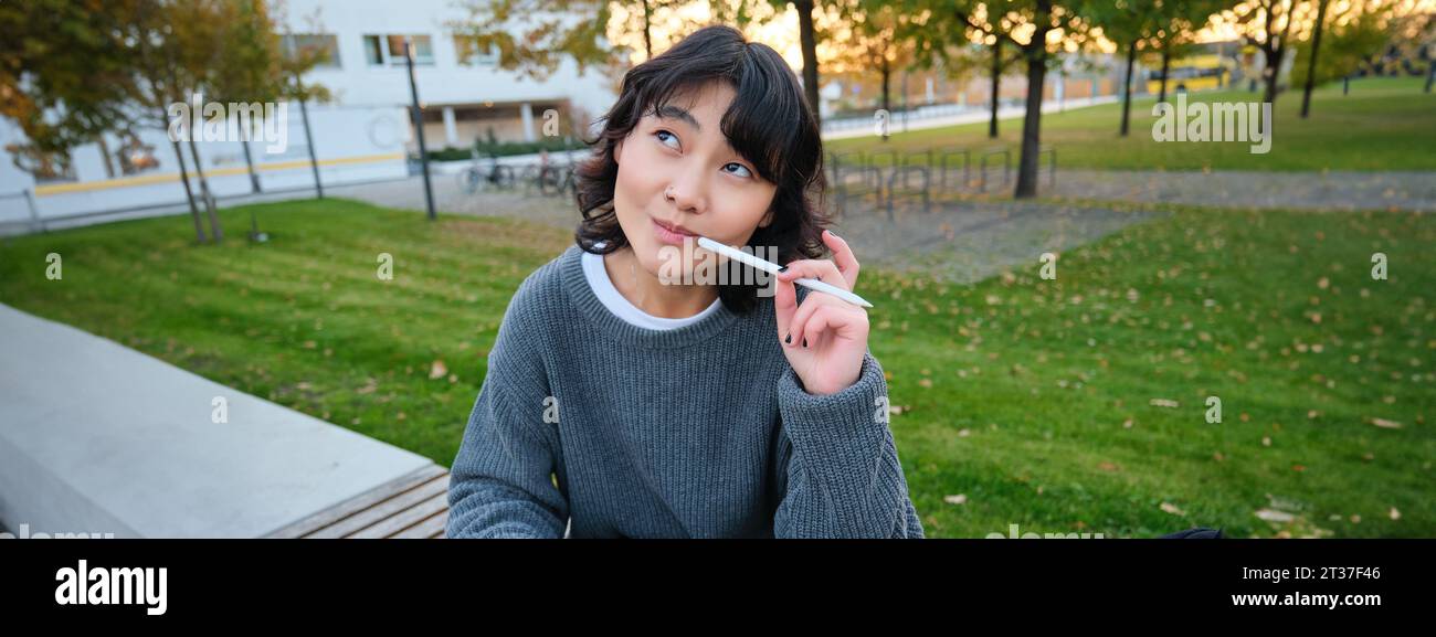 Portrait of asian woman, student in park, sits on bench with digital tablet and a pen, thinking, looking aside thoughtful, making notes, does her Stock Photo