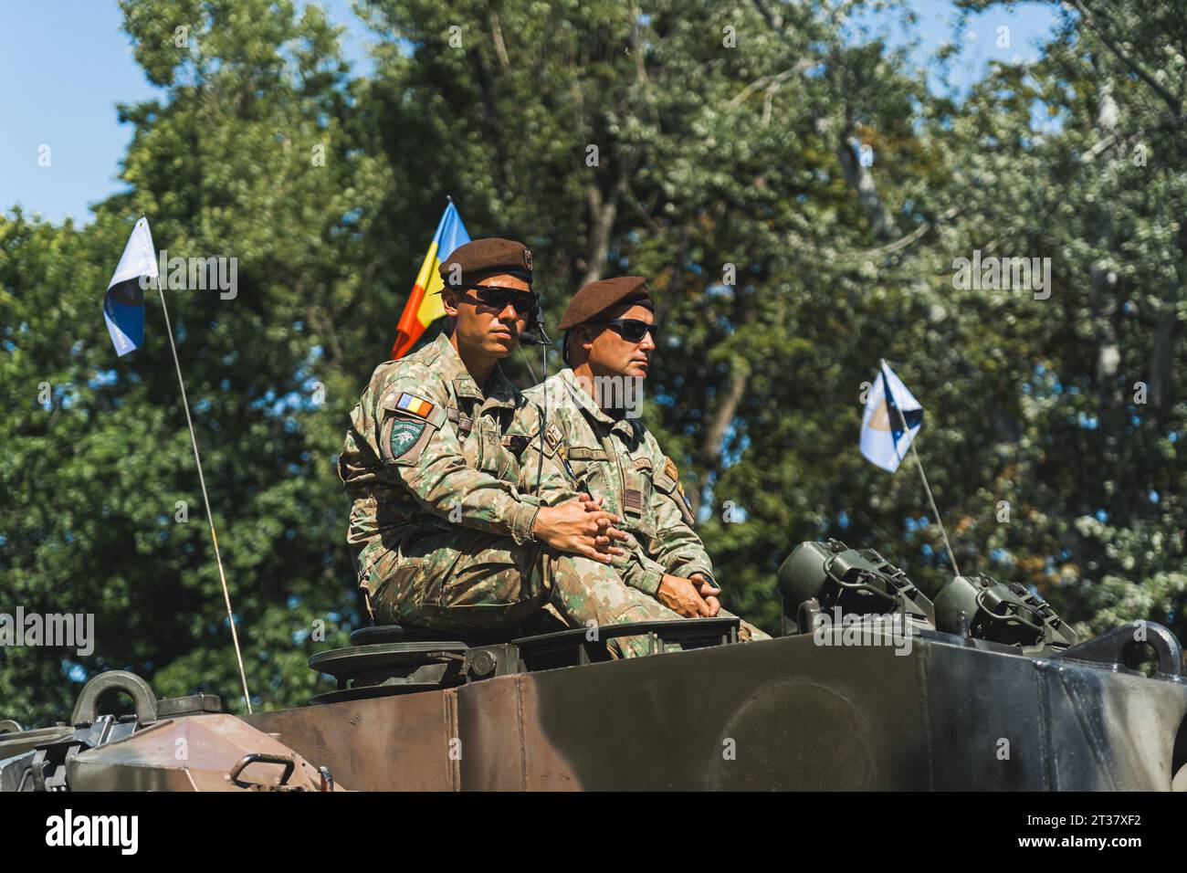 16.08.2023 Warsaw, Poland. Two relaxed Romanian soldiers in berets and sunglasses sitting on the top of the tank with Romanian flag and trees in the background. High quality photo Stock Photo