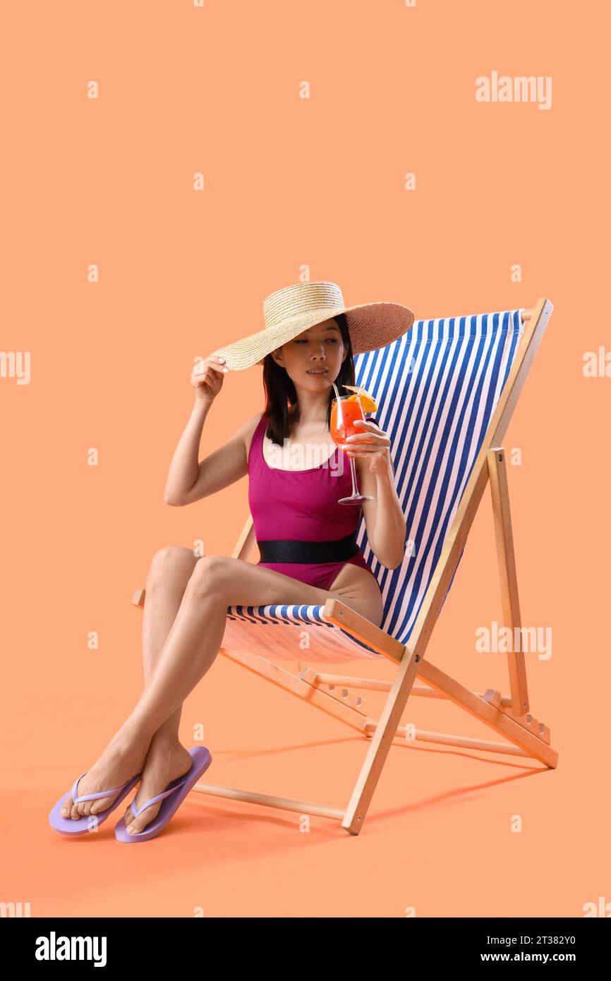 Beautiful young Asian woman in swimsuit with tasty aperol spritz sitting on deckchair against orange background Stock Photo