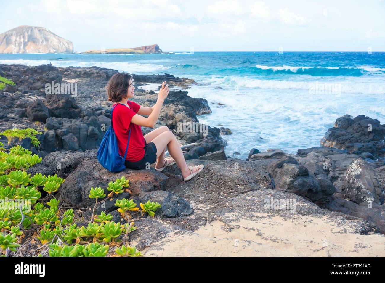 Young woman sitting on lavarocks along the Hawaiian ocean at Makapu'u beach taking pictures with cellphone Stock Photo