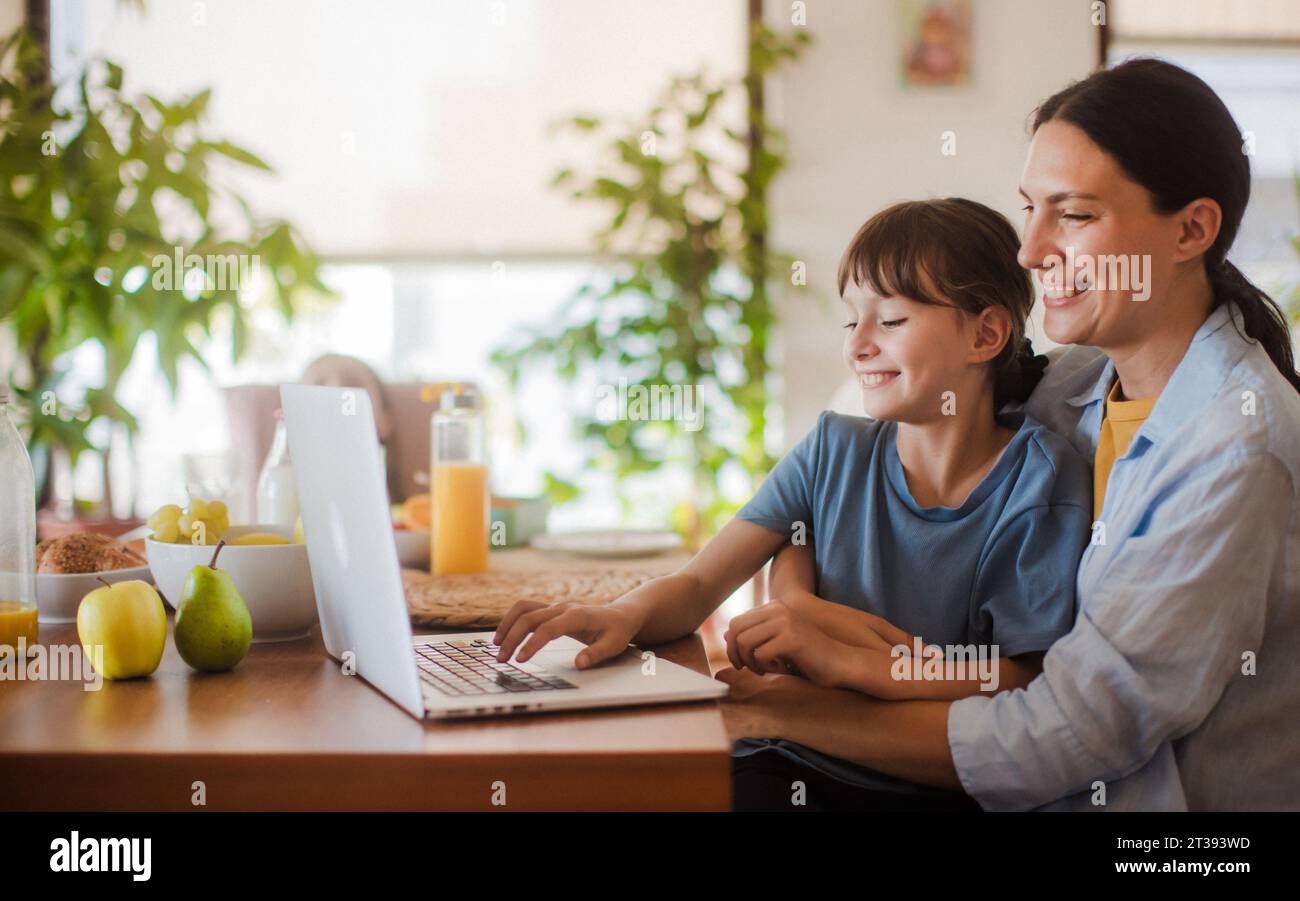 Mom and daughter watching entertaining videos on mom's work laptop. Remote work, home office for mothers with children. Stock Photo