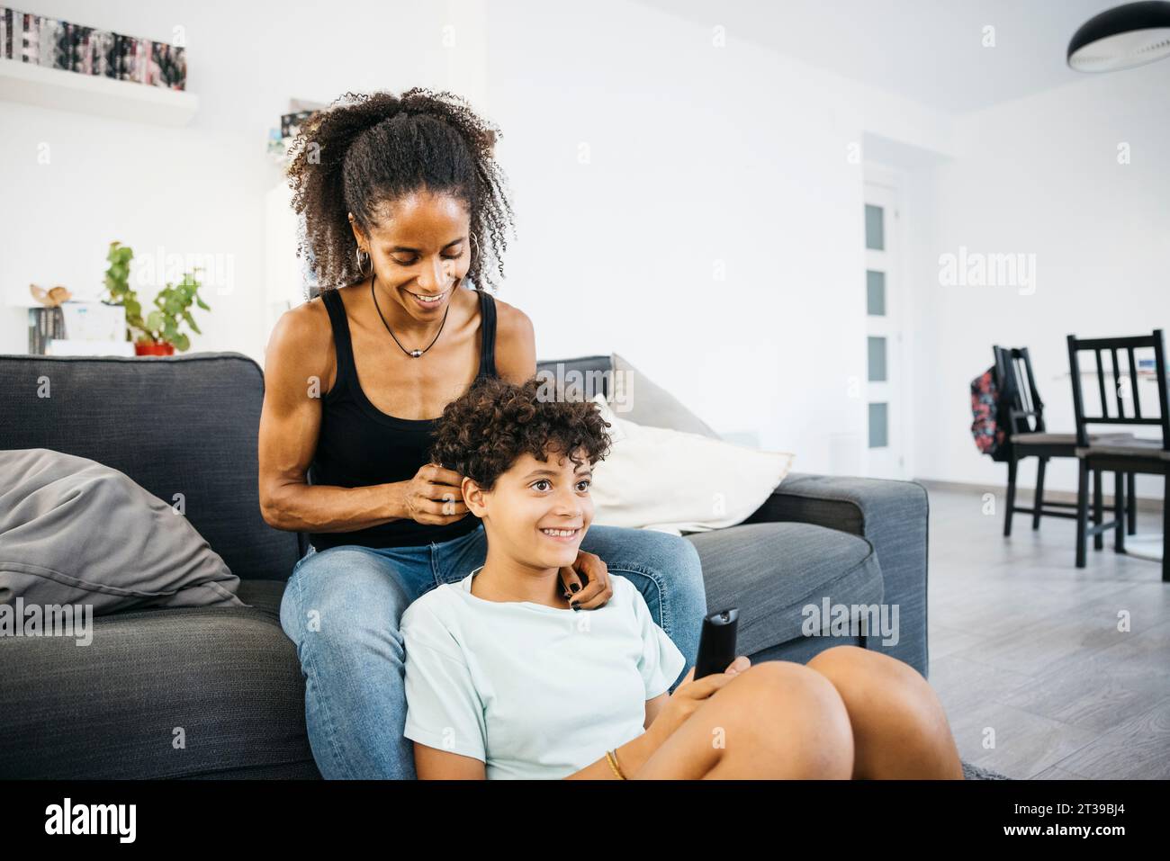 Young smiling mother combing her daughter on a sofa while watching tv. Mother and daugther spending time together infront of the tv. Stock Photo