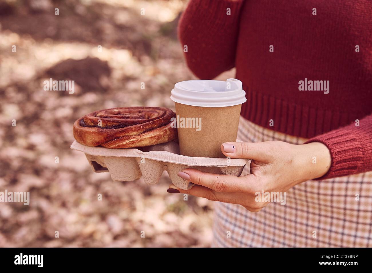 Woman walking in the park with cinnabon bun and coffee. Aesthetic autumn breakfast. Stock Photo