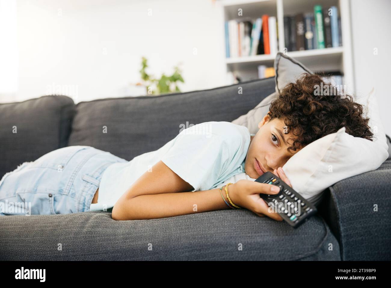 Ten years old bored girl lying on a sofa and watching tv, with a tv controller, in a living room. Girl watching a movie at home. Stock Photo