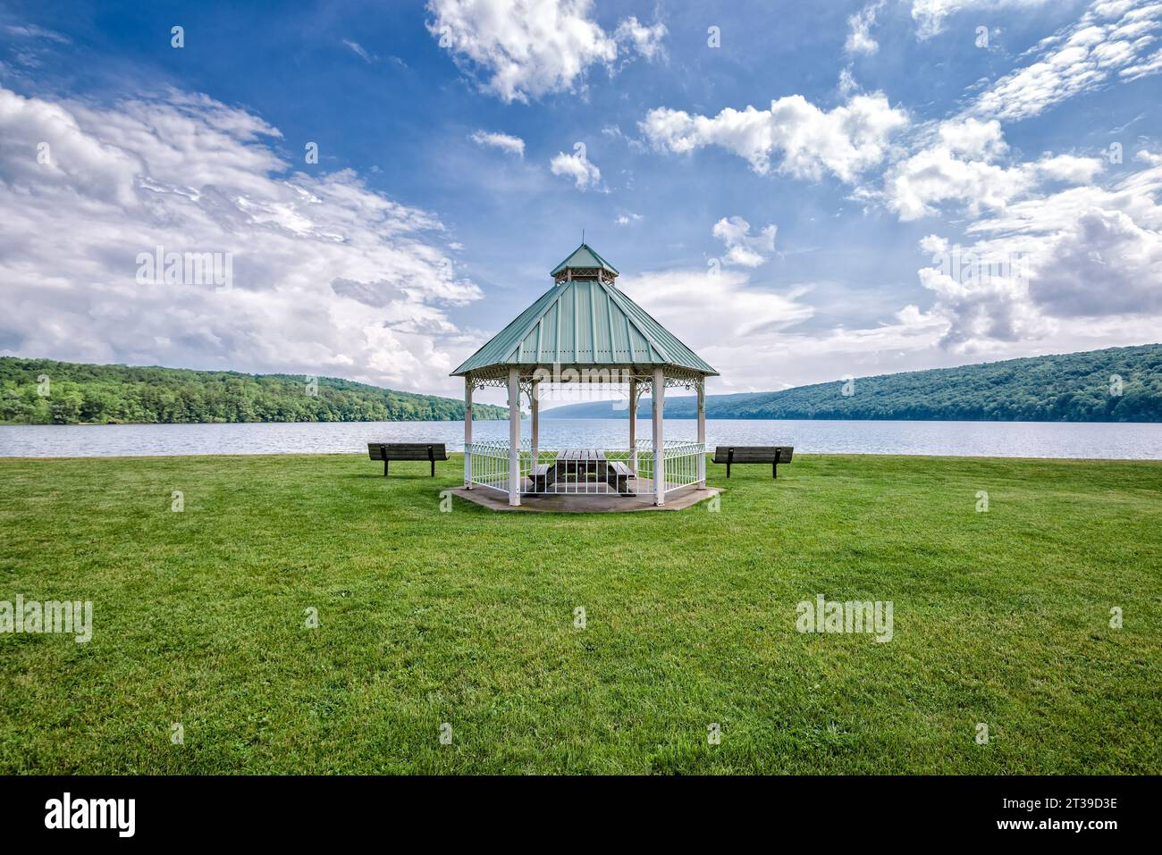 Picturesque view of drinking water lake with green grass lawn relaxing benches and lonely roof supported on pillars under cloudy blue sky in daylight Stock Photo