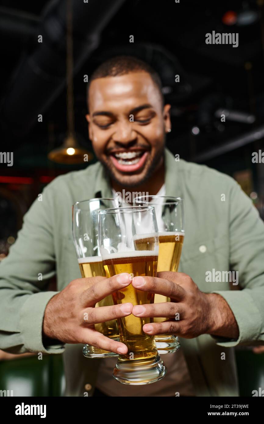 cheerful african american man holding three glasses of beer in bar, having fun time during weekends Stock Photo