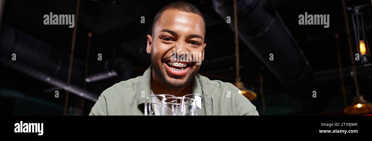 banner of happy african american man with braces holding three glasses in bar, having fun Stock Photo