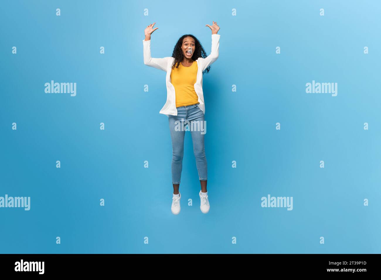 Full length portrait of smiling ecstatic African American woman jumping with arms raised in isolated light blue color studio background Stock Photo