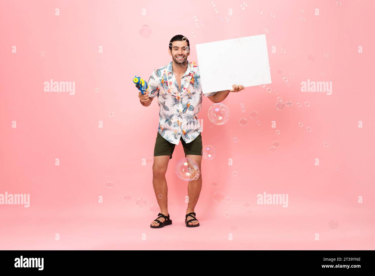Handsome smiling young Caucasian tourist man holding water gun and empty white board with space for text in studio pink background for Songkran festiv Stock Photo