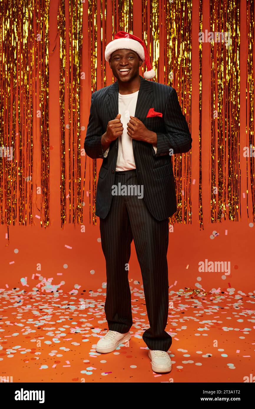 cheerful african american man in santa hat and black suit on orange backdrop with shiny decor Stock Photo
