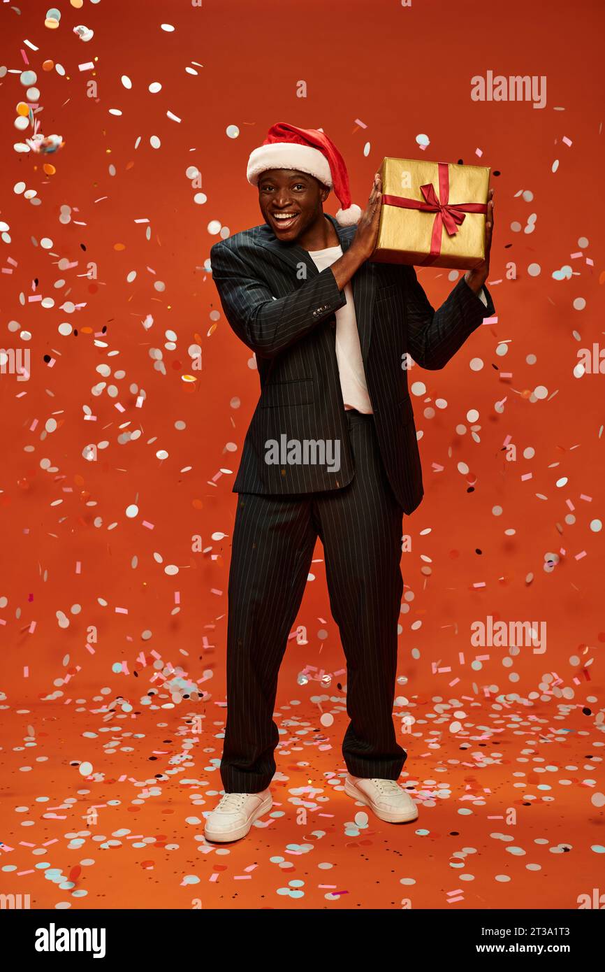 cheerful african american man in elegant suit and santa hat holding present under confetti on red Stock Photo
